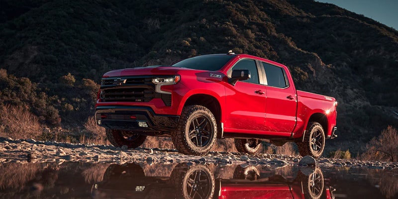 A red 2023 Chevy Silverado 1500 parked by a pond, reflecting in the water with mountains in the background.