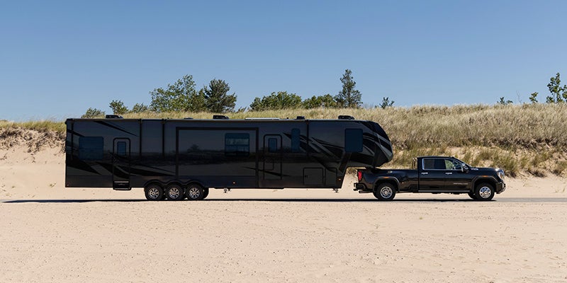 A black GMC Sierra HD pickup truck towing a large black trailer on a sandy area with trees in the background.