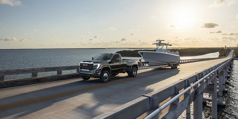 A black GMC Sierra HD pickup truck towing a white boat on a bridge during sunset.